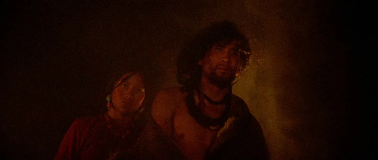 Horse Thief movie still: medium — A dark, dimly lit interior space with warm orange firelight glow. A man and woman stand side-by-side while looking…