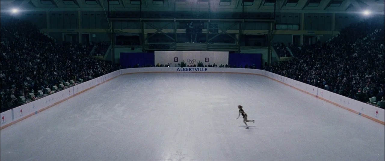 I, Tonya (2017) movie still: wide — A large indoor ice rink features banked seating filled with spectators under bright lights. A lone figure skater…