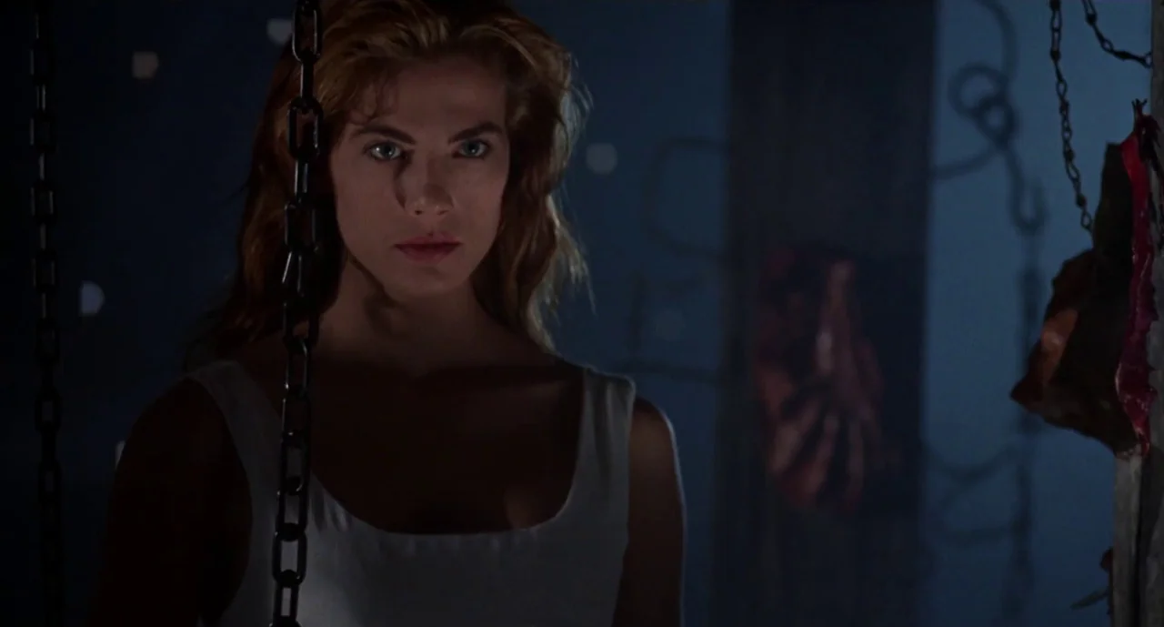 Hellraiser III: Hell on Earth movie still: medium — A dark, ominous room contains hanging metal chains and shadowy structural columns. A young woman…