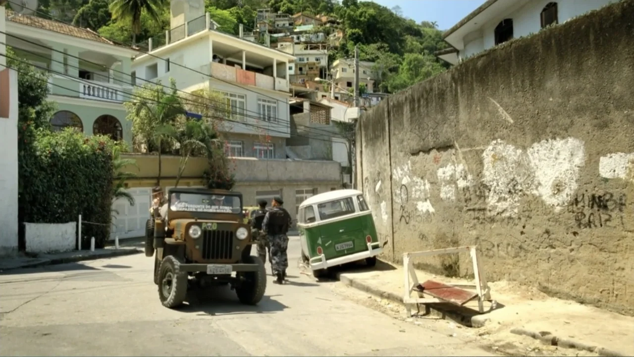 For Sale or Rent (2013) movie still: wide — A paved street runs between a textured concrete wall and white residential buildings. Officers in…
