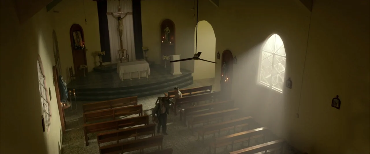 I Dream in Another Language (2017) movie still: wide — A quiet church interior features rows of wooden pews leading to a raised altar. Two men walk…