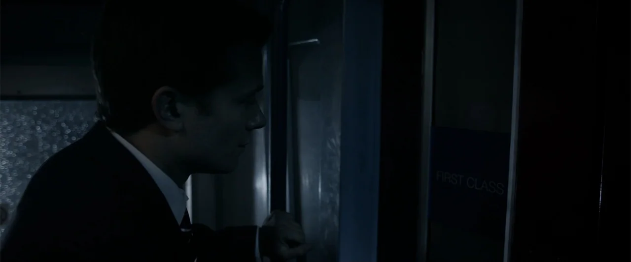 Howl (2015) movie still: medium — A dark airplane cabin interior with a door featuring a First Class sign. A man stands in profile wearing a suit…