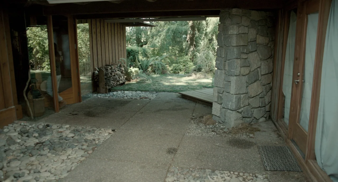 En lo profundo del bosque movie still: wide — A stone-paved patio connects a modern wooden structure to a lush green outdoor area. A stack of…