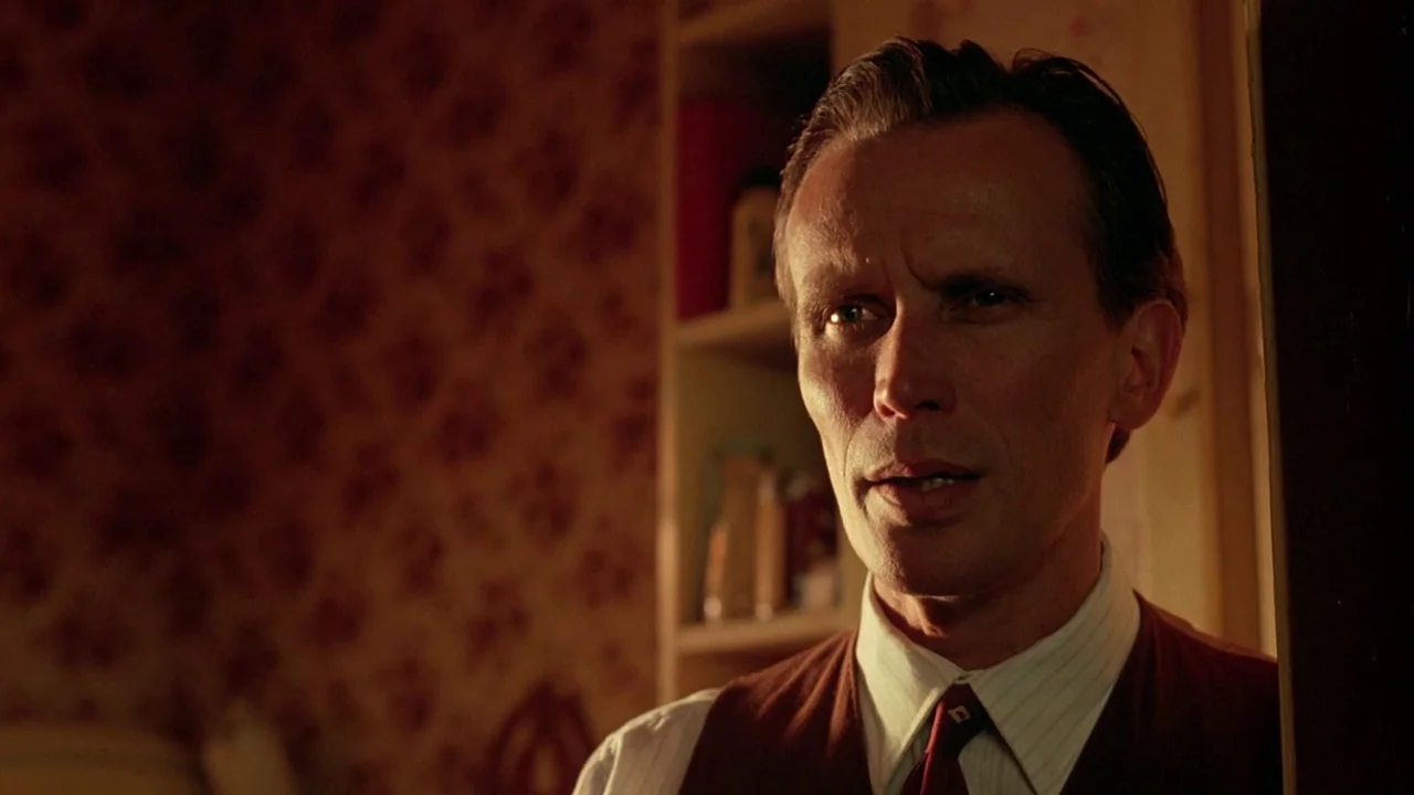 Naked Lunch (1991) movie still: closeup — A dimly lit room features dark wallpaper with a repeating floral pattern. A man in formal attire stands in…