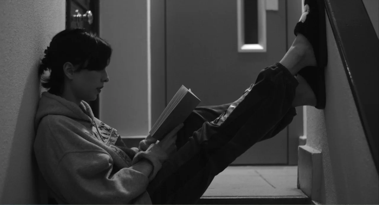El Planeta (2021) movie still: medium — A woman sits against a stairwell wall while reading a book held in her hands. She rests her feet against the…