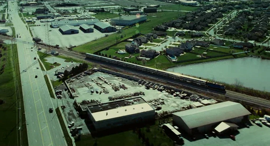 Source Code (2011) movie still: establishing — An aerial view shows a suburban area with warehouses, residential houses, and a pond. A long…