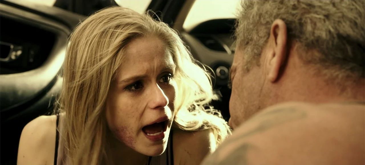Blood Father (2016) movie still: closeup — Two individuals sit inside the front of a vehicle during the daylight hours. A blonde woman speaks loudly…