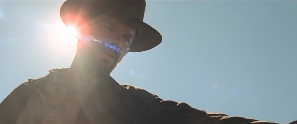 In A Valley of Violence (2016) movie still: medium — A man stands outdoors under a clear bright blue sky. Intense sunlight shines behind his hat…