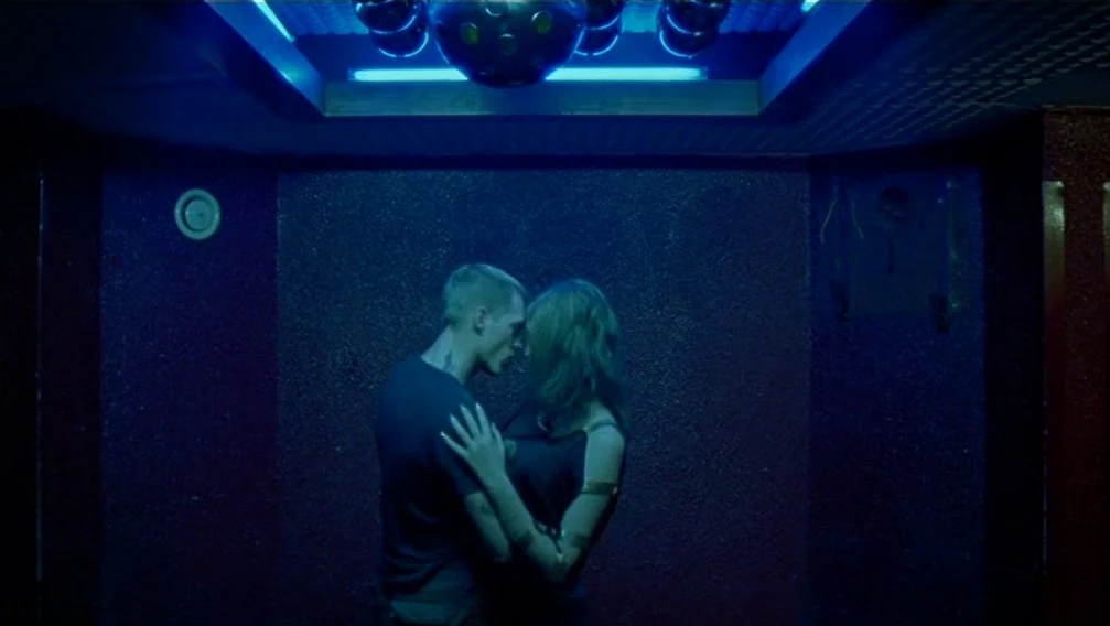 Import Export (2007) movie still: medium — A dark, windowless interior space features deep red, textured walls and blue overhead lighting. A man and…