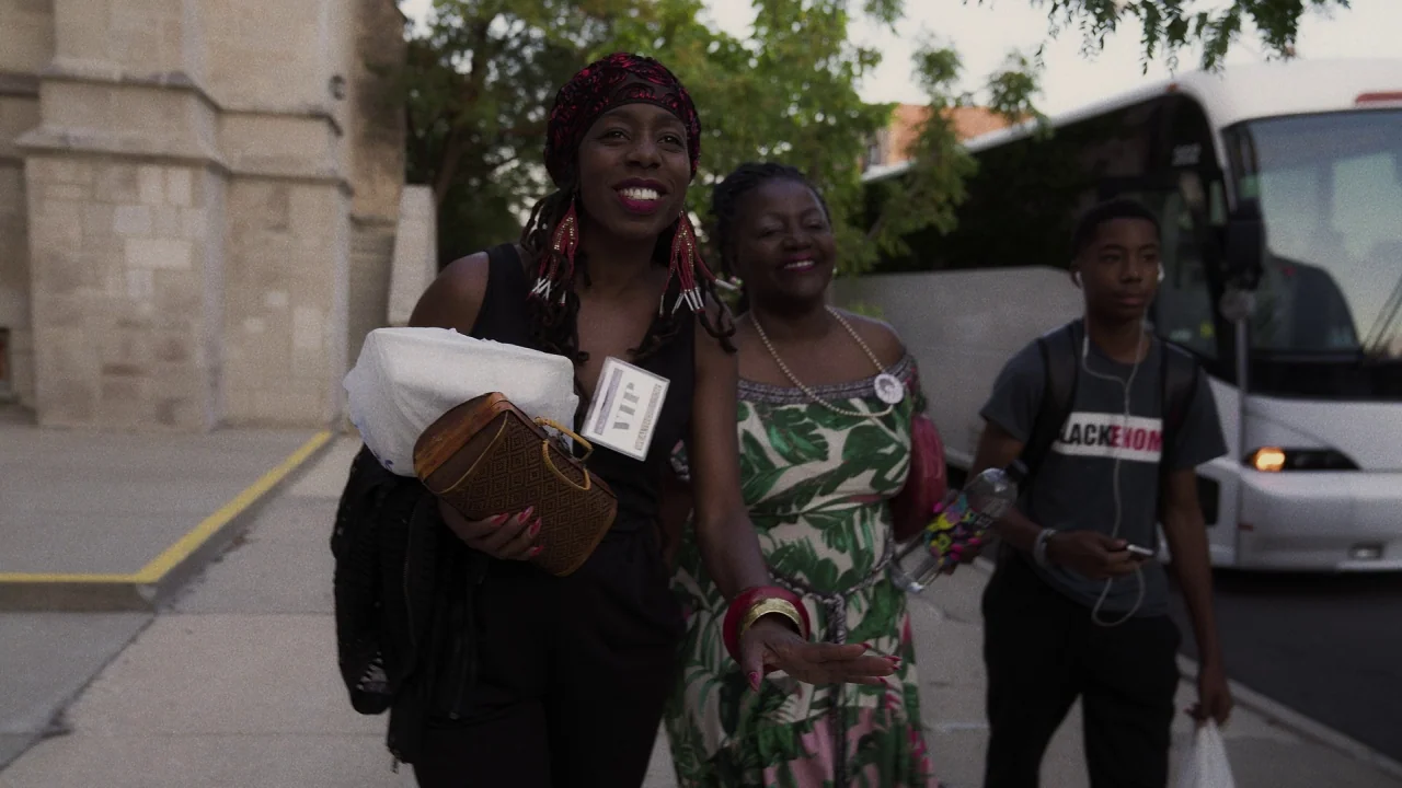Pass Over (2018) movie still: medium — A group of three people walk along a concrete sidewalk near a large parked bus. An stone architectural…