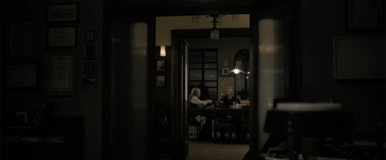The Equalizer 3 (2023) movie still: wide — A dimly lit room viewed through a doorway looking into an adjacent dining area. Two men sit at a table…