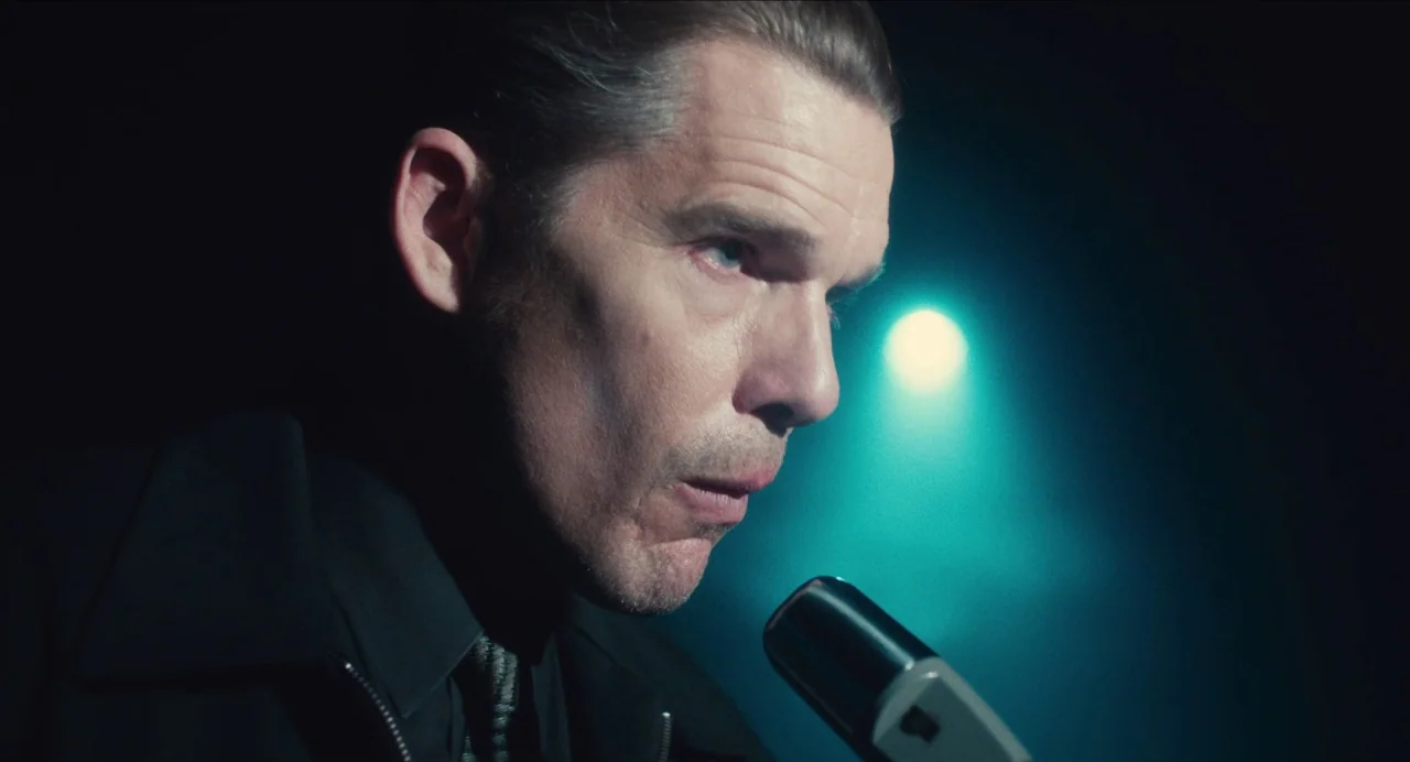 Born To Be Blue (2015) movie still: closeup — A dark space with a cyan light source behind the man. A man speaks into a vintage-style microphone at…