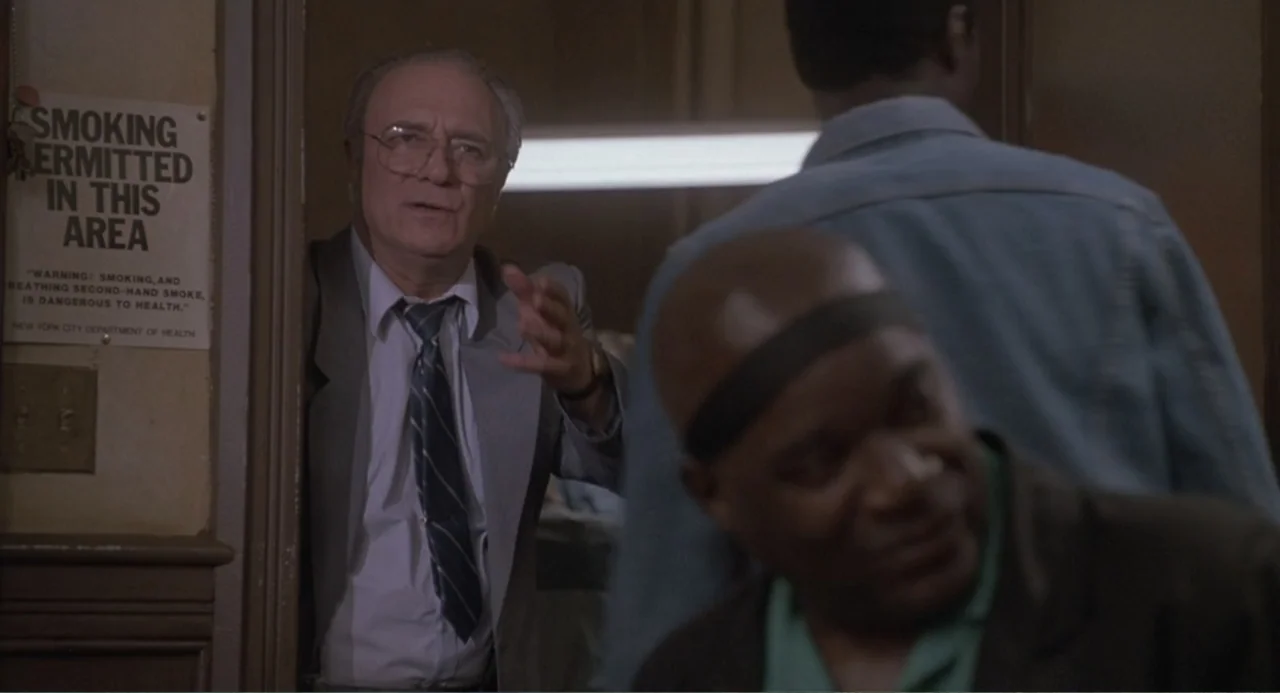 F/X2 (1991) movie still: medium — A dimly lit indoor space with a wall sign containing text. An older man stands in a doorway talking to a partially…