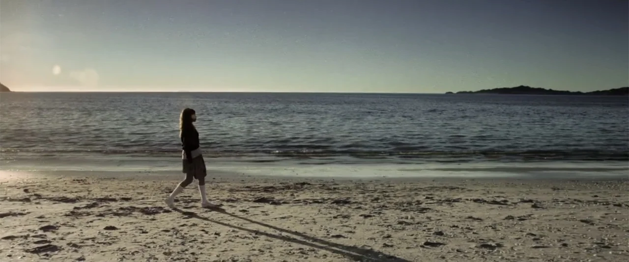 At Dawn (2013) movie still: fullbody — A wide shoreline features damp sand and calm ocean water under a clear sky. A young woman walks across the…