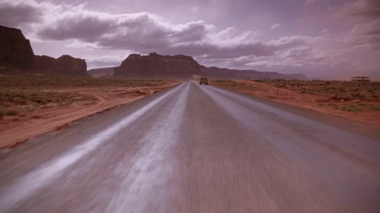 Natural Born Killers (1994) movie still: establishing — A long paved road stretches through a vast, arid desert landscape under a cloudy sky. A…