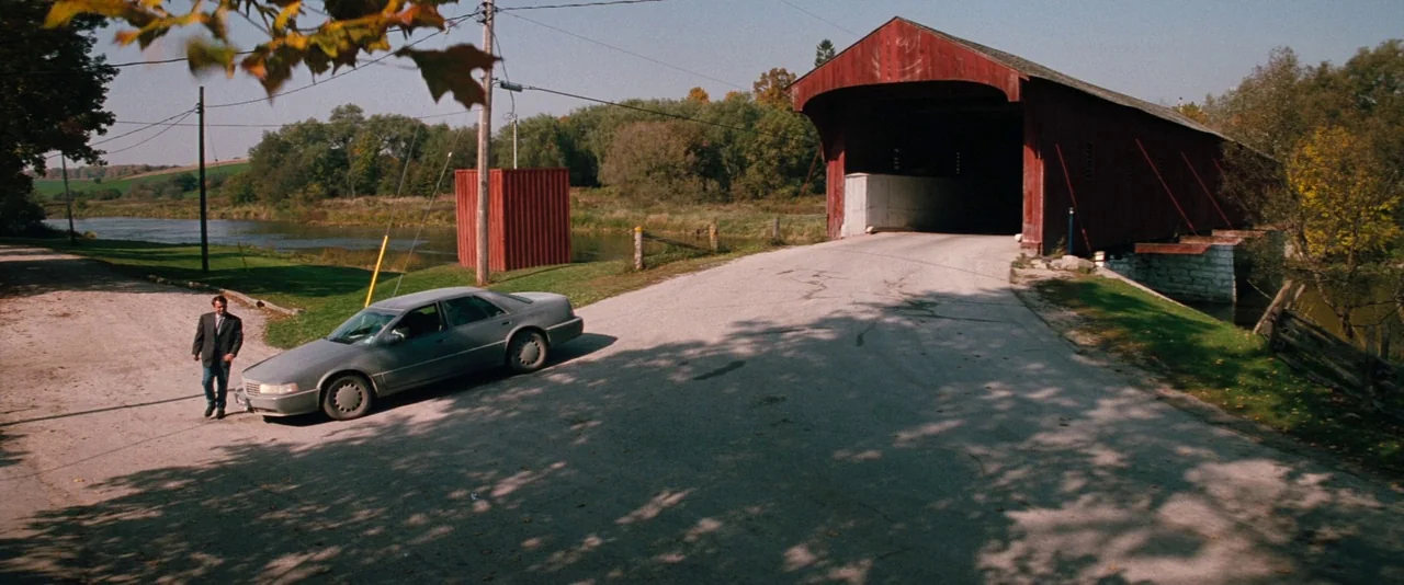 In The Mouth of Madness (1994) movie still: wide — A red wooden covered bridge spans a river next to a gravel turnout road. A man in a dark suit…