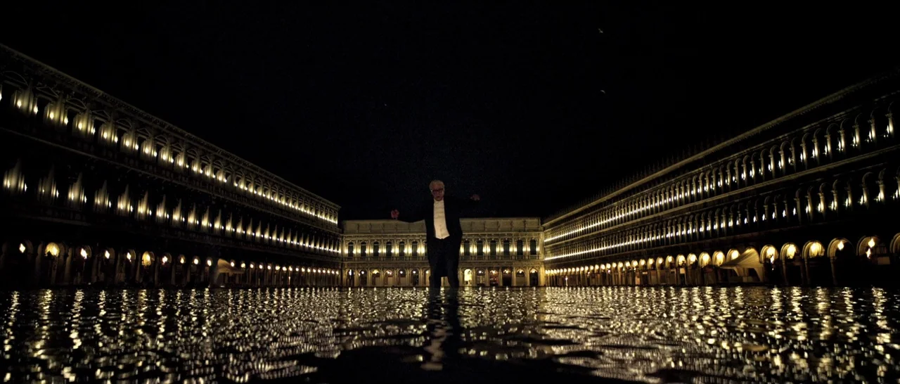Youth (2015) movie still: wide — St. Mark's Square in Venice is flooded at night with building reflections on water. A lone man in a suit stands in…