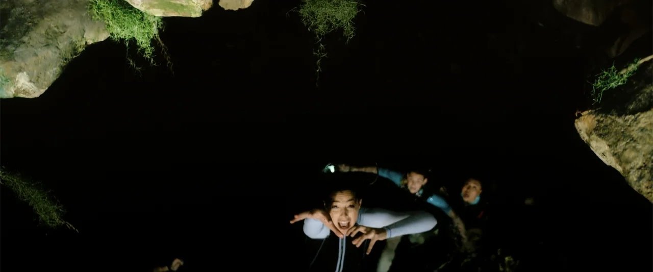 Anaconda (2024) movie still: medium — A dark underwater cave opening features moss hanging from the jagged limestone ceiling above. Three divers in…