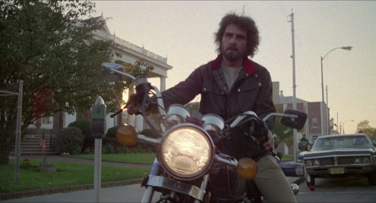 The Amityville Horror (1979) movie still: medium — A residential street lined with houses and parked vintage cars during the golden hour. A man…