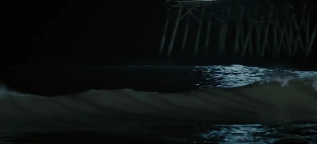 Merv (2025) movie still: establishing — A wooden pier structure stands against a dark night sky above dark water. Ocean waves break in the…