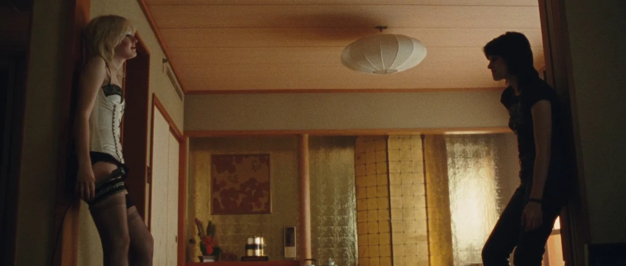 The Runaways (2010) movie still: wide — A hotel room with wood-paneled walls and a circular pendant lamp hanging overhead. One woman in a corset…