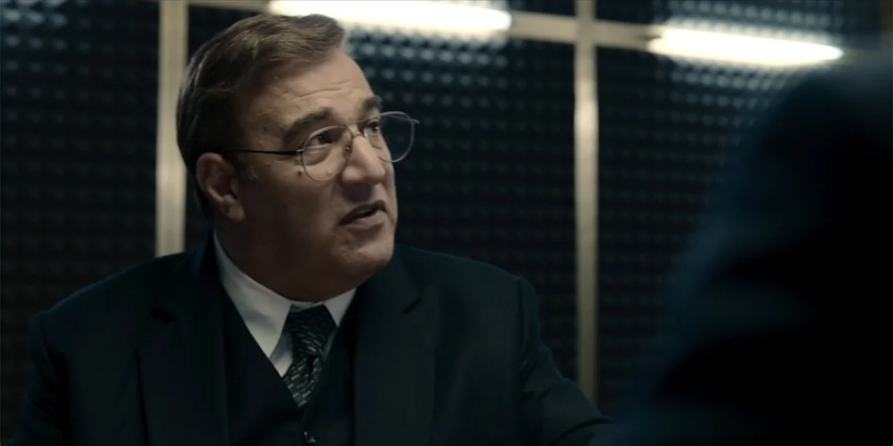 Oslo (2021) movie still: medium — An interrogation room features dark acoustic panel walls with silver vertical metal supports. A man in a suit sits…