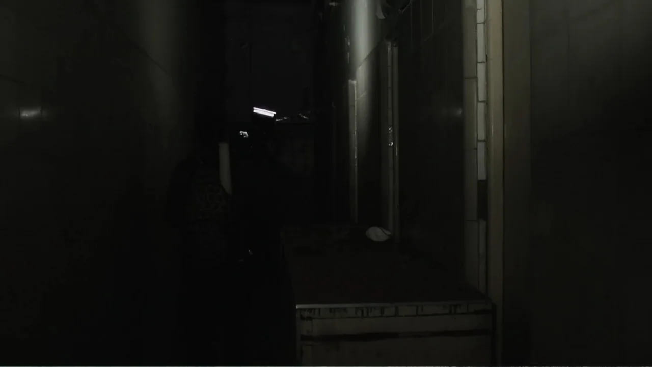 Lost (2010) movie still: medium — A dark, narrow corridor features tiled walls and a metal surface in low light. A person stands facing away from…