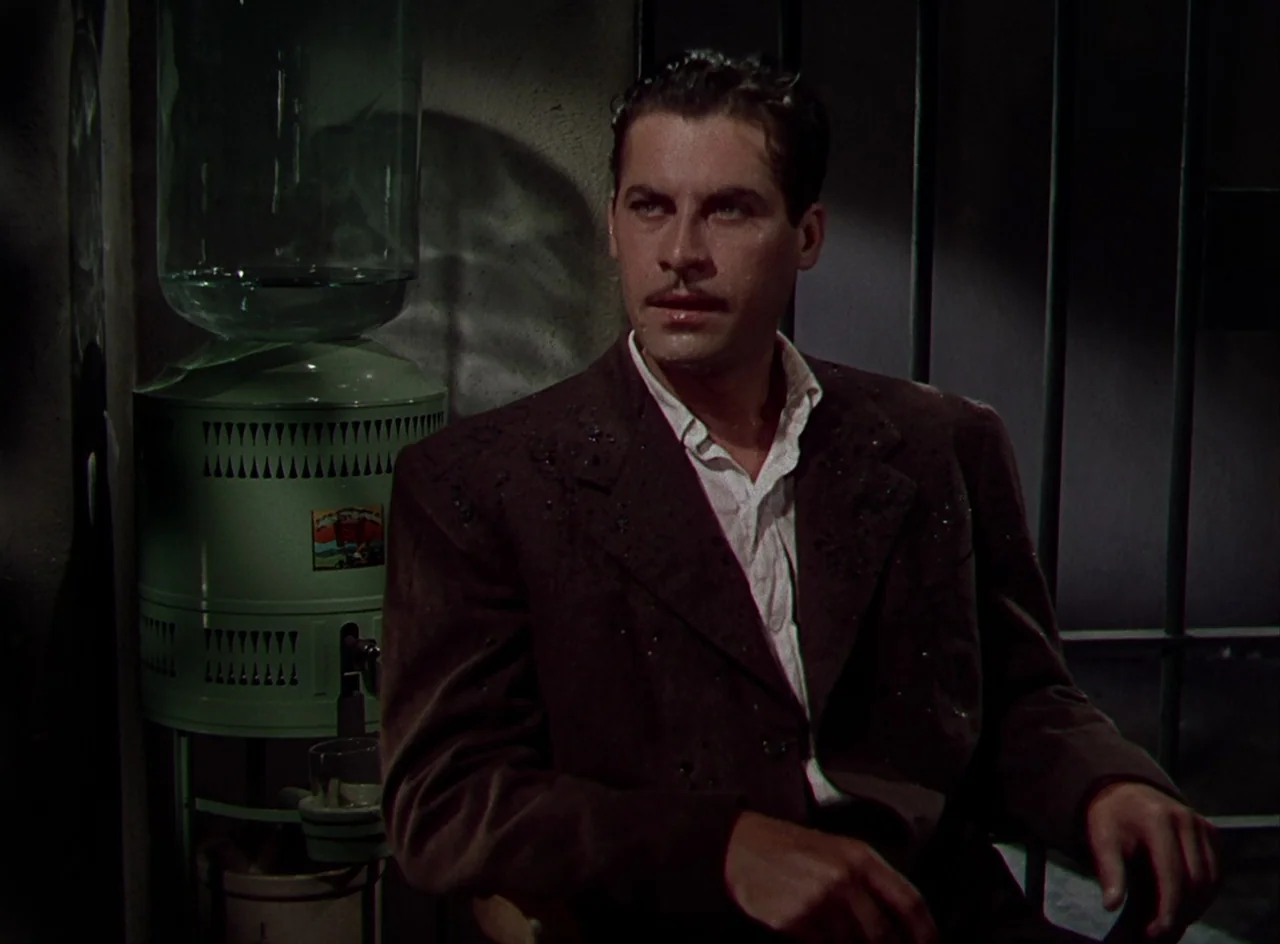 Desert Fury (1947) movie still: medium — A dimly lit room with stone walls and vertical metal bars behind the man. A green vintage water dispenser…