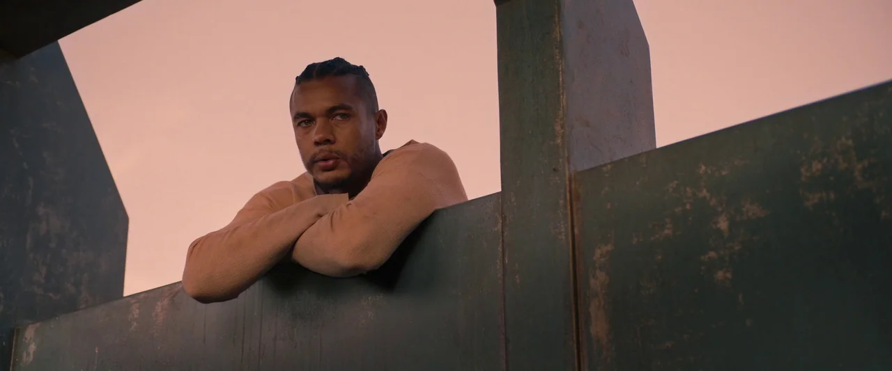 Settlers (2021) movie still: medium — A rooftop structure features weathered green metal panels against a clear sunset sky. A young man with braids…