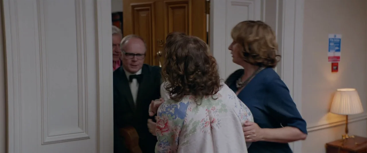 Hampstead (2017) movie still: medium — A hotel hallway with off-white walls features a partially open doorway. Two women stand in the corridor…