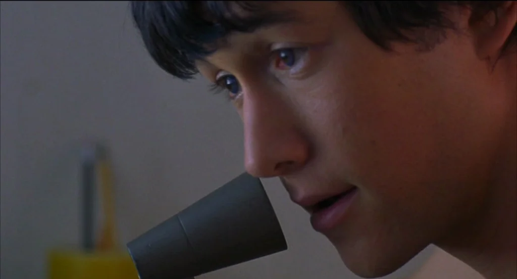 Mysterious Skin (2004) movie still: closeup — A close-up shot of a young man speaking into a phone receiver. He is positioned against a plain, dimly…