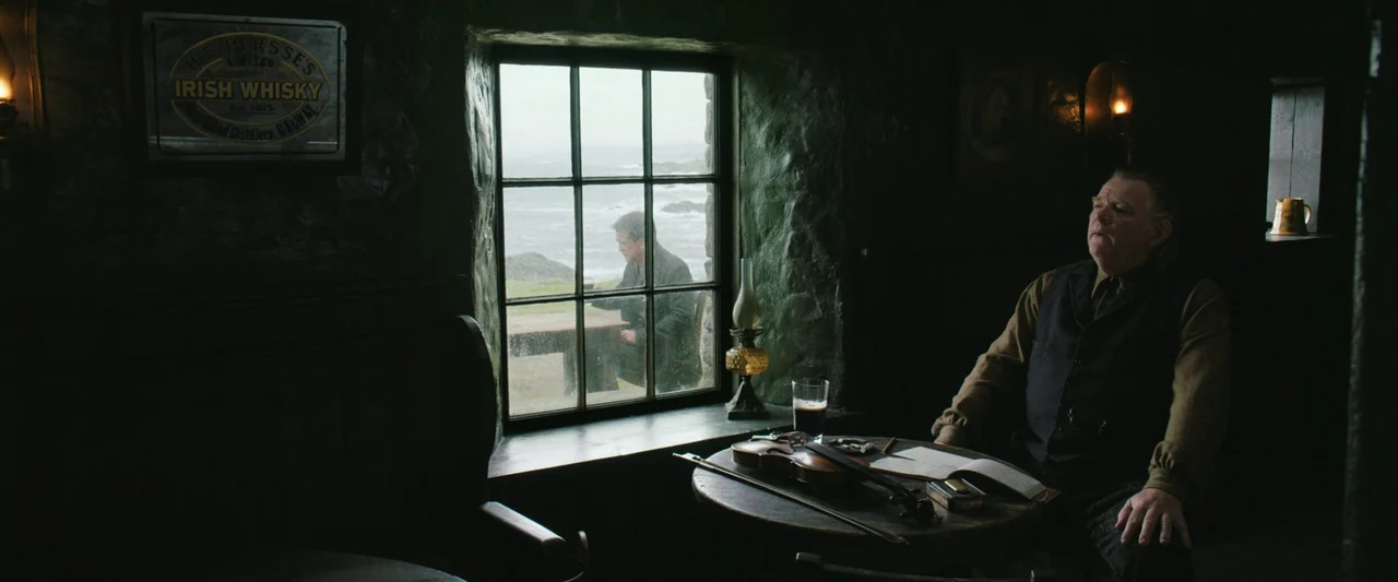The Banshees of Inisherin (2022) movie still: medium — An old, dark Irish pub interior features heavy stone walls and small oil lamps. A man sits at…