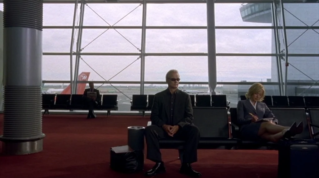 Broken Flowers movie still: wide — An airport departure lounge features large floor-to-ceiling glass walls overlooking a landing strip. A man…