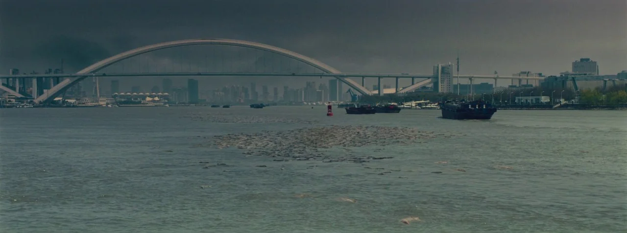 Dead Pigs (2018) movie still: establishing — A wide river flows beneath a large arched bridge near a modern urban skyline. Hundreds of dead pig…