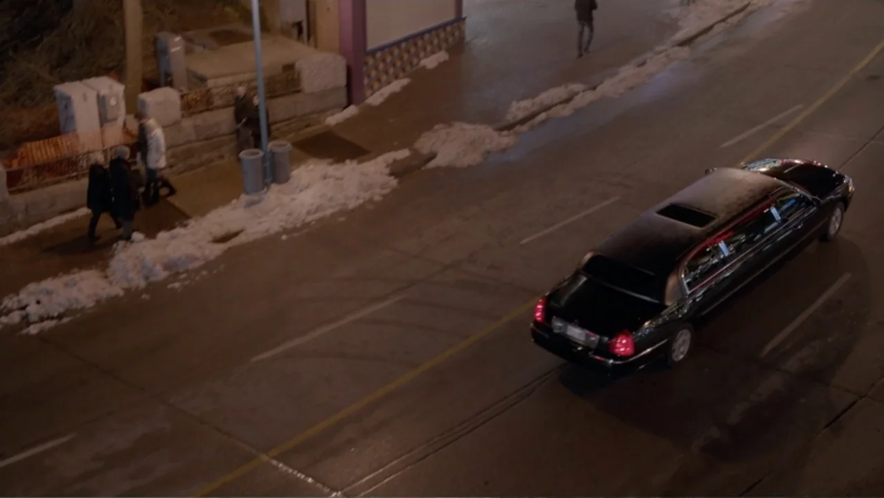 The Captive (2014) movie still: wide — An asphalt road features a black limousine moving right while snow lines the sidewalk. Pedestrians walk along…