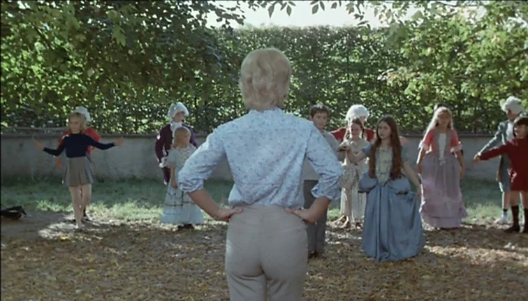 Le Boucher (1970) movie still: fullbody — A group of children and an instructor stand in a circle in a leafy park. The instructor stands with her…