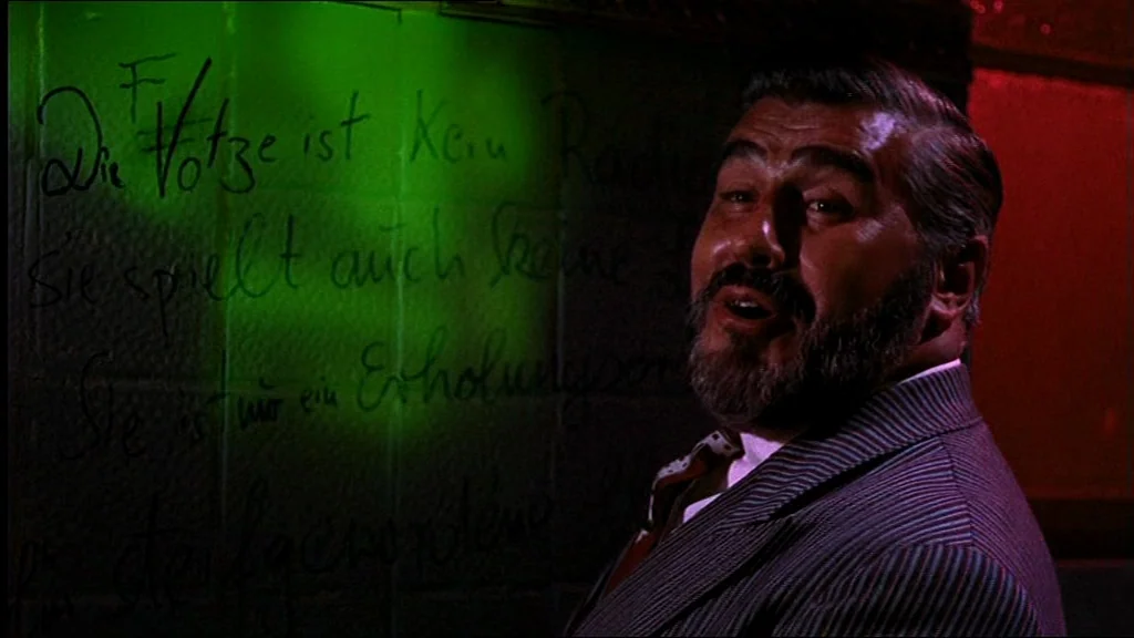 Lola (1981) movie still: medium — A dimly lit bar wall features German text written in black marker. A man in a striped suit jacket stands in front…