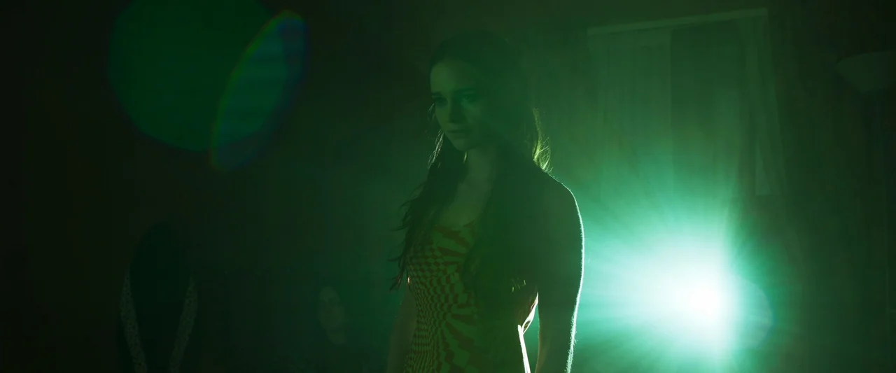 Stopmotion (2024) movie still: medium — A dimly lit interior space features a young woman standing in green light. A bright lens flare from a light…