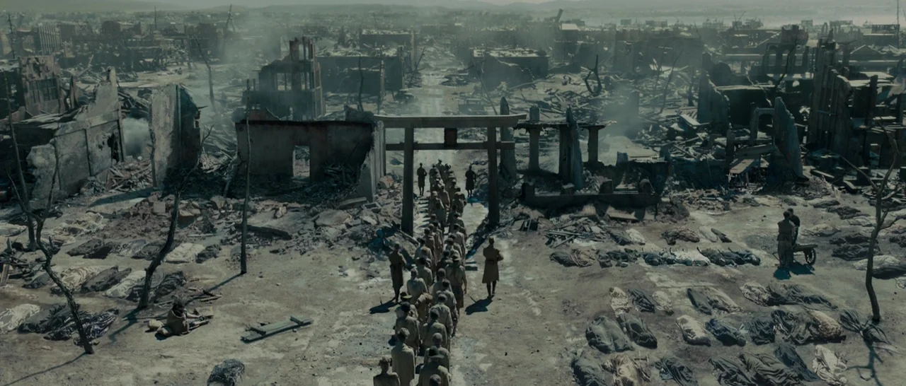 Unbroken (2014) movie still: establishing — A decimated city landscape is filled with rubble and rows of covered bodies. A line of soldiers walks…