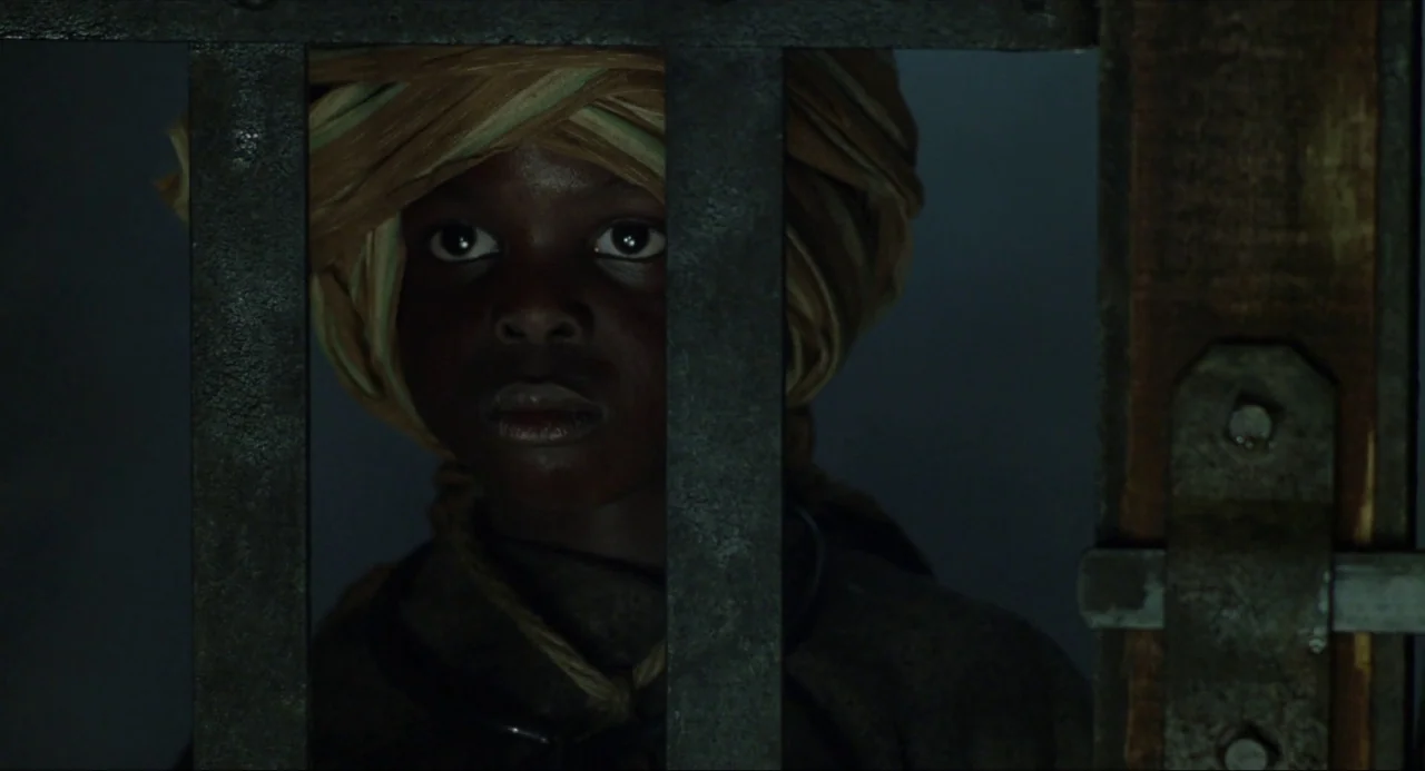 Amistad (1997) movie still: closeup — A dark, dimly lit prison cell features heavy vertical metal bars. A young boy wearing a patterned headwrap…