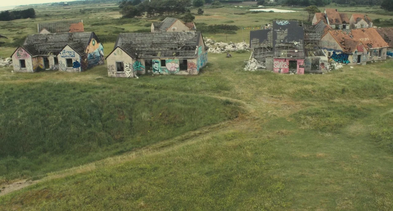 Faces Places (2017) movie still: establishing — A cluster of derelict single-story houses with damaged roofs sits in an open field. The buildings…