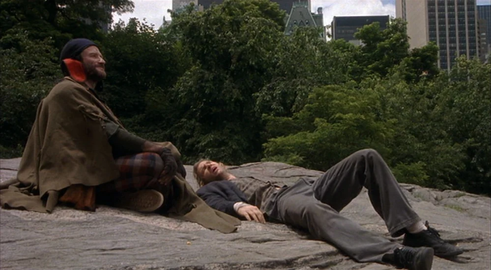The Fisher King (1991) movie still: fullbody — A flat rocky outcrop in a city park is surrounded by dense trees and buildings. Two men rest together…