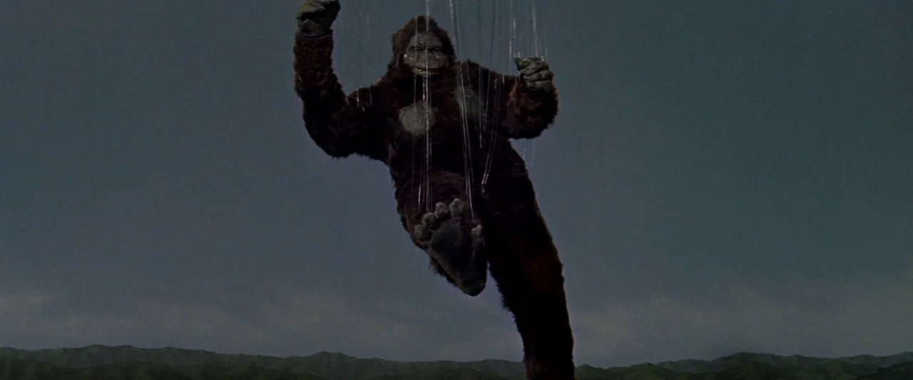 King Kong Vs. Godzilla (1962) movie still: fullbody — A dark, fur-covered ape figure is suspended against a dull, cloudy sky. Multiple thin wires…