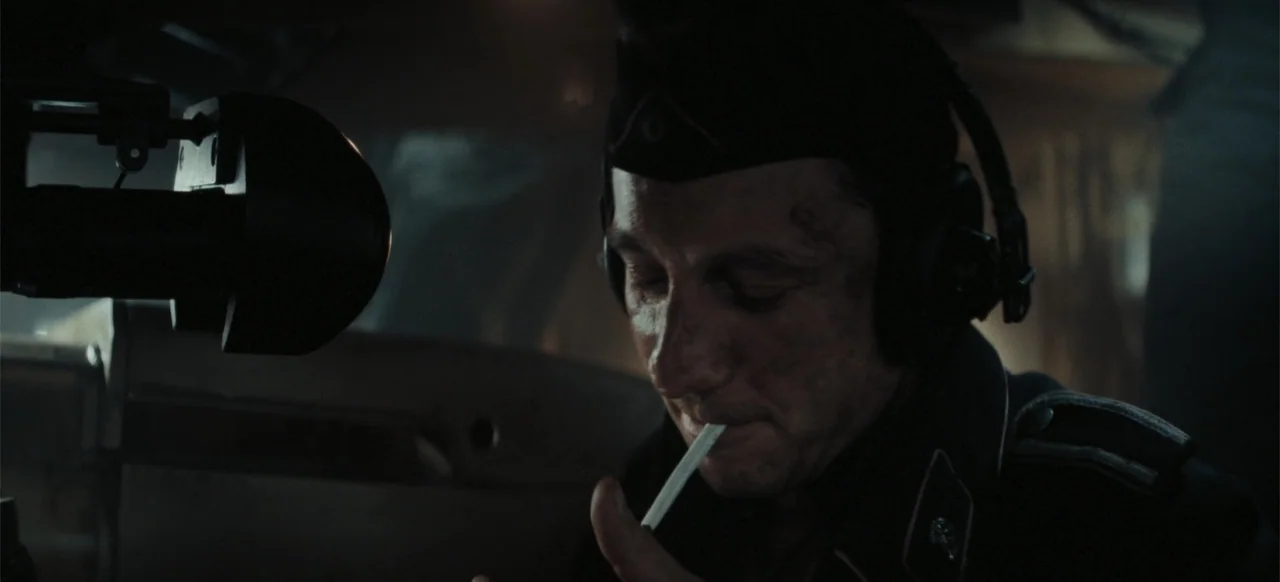 The Tank (2025) movie still: closeup — A dimly lit cockpit with mechanical panels visible in the dark background. A man in a dark military uniform…