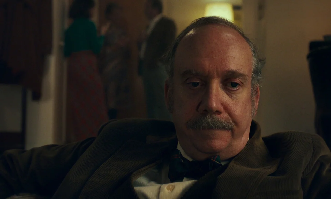 The Holdovers (2023) movie still: closeup — A dimly lit room provides a backdrop for a man in formal wear. He sits in the foreground while blurred…