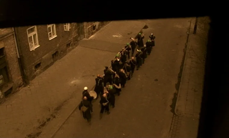 Golem (1980) movie still: wide — A narrow cobblestone urban thoroughfare at night is flanked by brick buildings. A small group of musicians in dark…