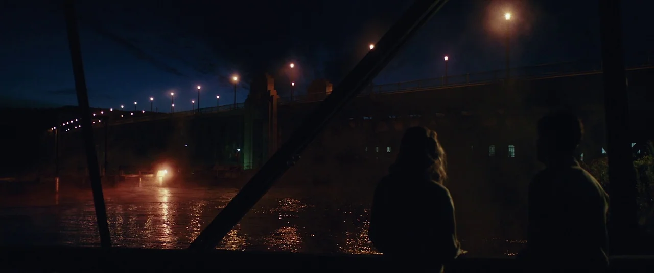 The Man Who Killed Hitler and Then the Bigfoot (2018) movie still: wide — A night view across a river toward a long bridge lined with glowing…