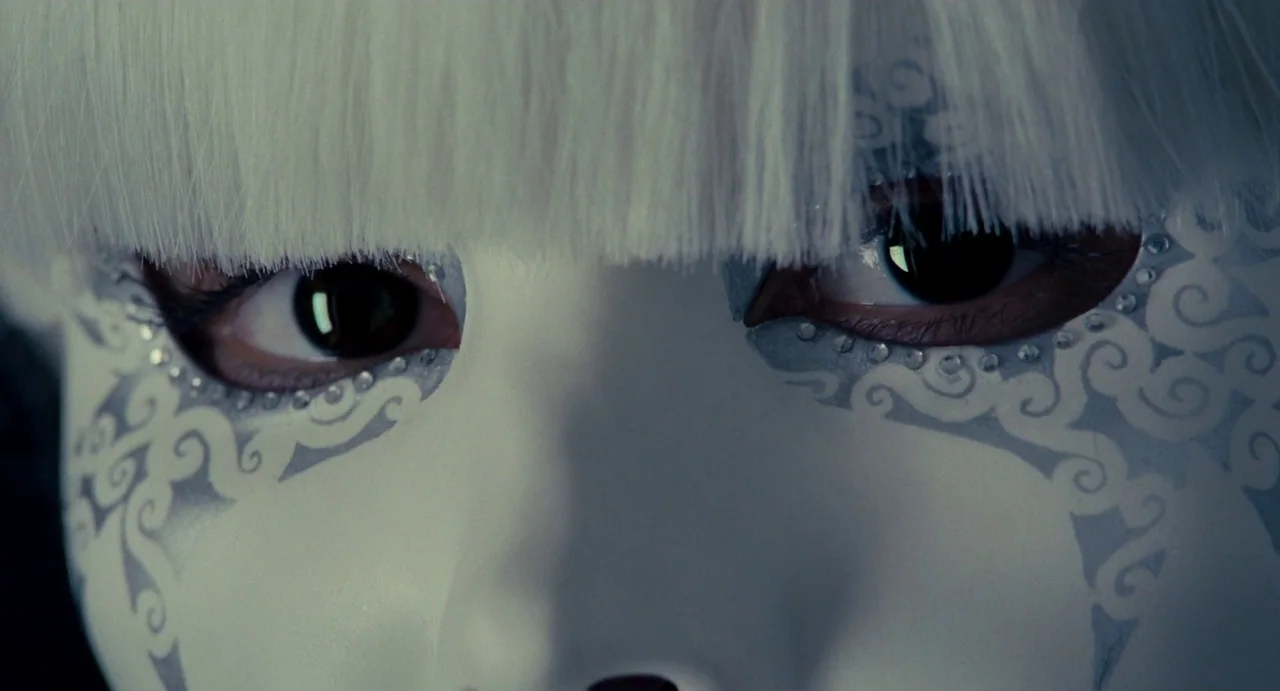 Passion (2012) movie still: closeup — Close-up of a decorative white face mask with silver filigree patterns and sequins. Two dark eyes look through…