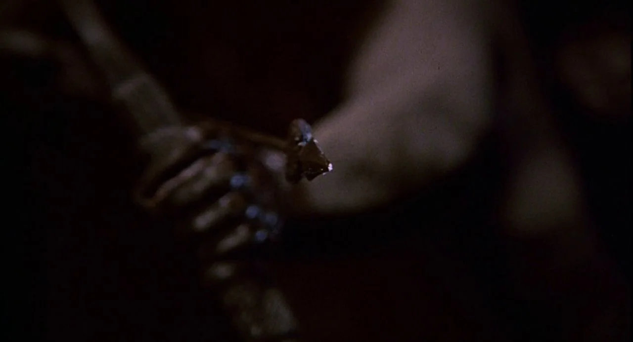 Psycho III (1986) movie still: closeup — A dark, undifferentiated interior space obscures all background detail in deep shadow. A metallic sword…