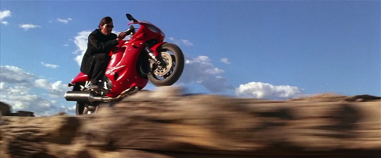 Mission: Impossible II (2000) movie still: fullbody — A dry, rocky desert landscape stretches under a bright blue sky with scattered clouds. A man…